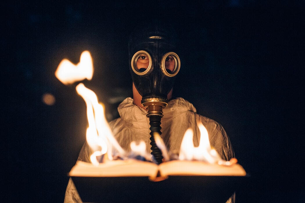 Person wearing a gasmask viewing a burning book on a black backdrop