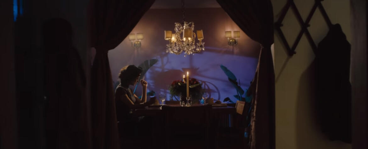 Wide shot looking into a room where a person is sitting at a dinner table with a chandelier and lit candles in the center.